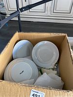 Box containing several white ceramic plates stacked with one upright showing bottom logo