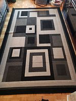 Photo of a 5x9 feet area rug with a geometric square and rectangular pattern in black, gray, and cream tones, laid out on a parquet floor between furniture.
