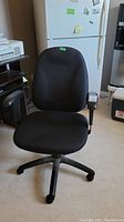 Front view of the black office chair placed indoors near a refrigerator and printer.
