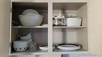 Wide view of cupboard with white casserole dishes, bowls, plates, glassware and white ceramic gravy boat.