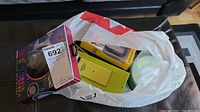 Photo taken showing the lot inside a white plastic bag on a black leather sofa, containing boxed items and loose accessories including a circular Bluetooth speaker in a colorful box and a yellow-green box likely for the fitness tracker.