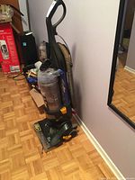 Upright Eureka vacuum with clear dust container, black and grey body, yellow latch, standing on parquet floor near wall and mirror.