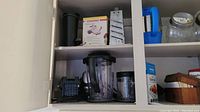 Kitchen cabinet shelf showing black manual slicer, grater, blender pitcher, and smaller blender container parts.