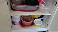 Photo showing an open kitchen cabinet with shelves containing various plastic food storage containers in different sizes and colors.