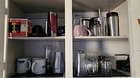 Upper shelves of cupboard showing boxed holiday mug, ceramic and insulated mugs, and assorted glasses and cups on lower shelves.
