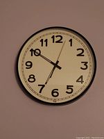 Front view of a round wall clock with white face, black numbers and black frame, showing time at about 6:50:03.