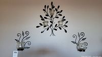 Wide shot of three metal wall candle holders mounted on a beige wall. The central holder has three glass votive cups; the two side holders each have one glass cup. Metal designs feature leaves and swirls.