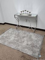 Area rug laid flat on floor with metal frame glass top side table placed above it. Rug has a silver two tone color and subtle pattern.