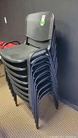 Stacked group of 8 black plastic chairs with metal frames against a wall, showing the shape and structure of the seat and backrest.