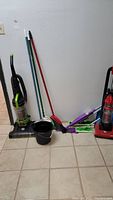 Photo of two upright vacuums, three brooms, a mop, and a black bucket arranged against a wall on tile floor.