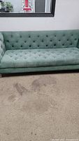 Full frontal view showing green button tufted upholstery on seat and backrest, wood-look frame and block legs, couch sitting on carpeted floor with visible dirt spots