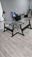 Wide angle photo showing the crescent shape glass top desk with black metal frame, black mesh office chair with wheels behind desk, and lamp on desk corner.