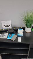 Full view of the shelving unit's top surface with items: faux plant in pot, Anker USB charging display stand, black power bank, electronics boxes, power bar.
