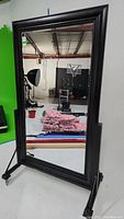Front view of the large rectangular floor mirror on a rolling metal stand showing the black frame and reflections of the studio background.