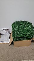 Overall photo showing faux greenery panels stacked in a box beside a sealed box of peel and stick floor tiles.