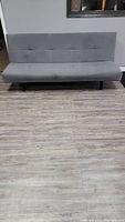 Gray futon couch placed against gray wall and light wood laminate flooring, viewed from front.