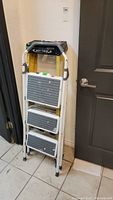 Full view of the folded step ladder showing three grey steps with paint splatters and white metal frame with grey padding.