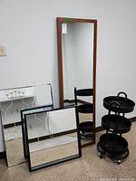 Three mirrors leaning against a white wall with a round black metal accessories cart on wheels beside them on carpeted floor.