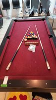 Full view of pool table with red felt, balls in triangle rack, two cues, cue rack and chalk on the surface