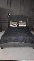 Front view of queen upholstered bed made with black bedding and two black and white striped pillows against dark wall