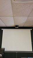 White pull-down projector screen mounted on a wall beneath ceiling tiles