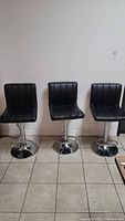 Three matching black adjustable bar stools with chrome pedestal bases and footrests, arranged side by side against wall