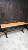 Wide angle photo showing full view of rectangular office table with light brown melamine top and black legs against black wall.
