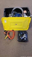 Yellow-lid black plastic tub filled with various tools and accessories, with extension cord and drills placed outside it on the floor.
