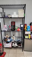 Full view of wire shelf loaded with miscellaneous items including paper products, coffee maker, cleaning supplies, and kitchen items, alongside a small fridge.