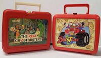 Front view of two vintage red plastic lunchboxes: The Real Ghostbusters and DuckTales with colorful stickers on front.