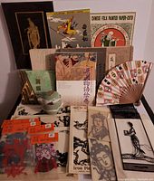 All lot items arranged on a table including paper cuttings, metal wall art, travel tea set, hand fan, and art catalogs