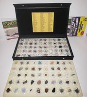Black display case open showing 100 individual circular compartments with various rock and mineral samples inside. Two educational boxes positioned behind the case.