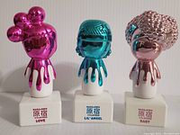 Front view of three metallic character-shaped perfume bottles sitting on white square bases labeled Love, Lil' Angel, and Baby.