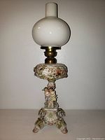 Full front view of the antique oil lamp featuring the milk glass chimney, brass burner, floral font reservoir, and cherub base.