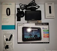 All four devices including the Fitbit Alta box and extra wristband, Garmin GPS with mount and power cord, Homder voice recorder boxed, and Supersonic tablet in box with cables.