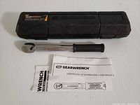 GearWrench branded black hard plastic case, torque wrench with black handle grip and silver shaft, instruction manual and calibration certificate laid out on a table.