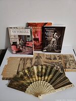 Photo showing three Victorian themed books standing upright behind vintage newspaper clippings and a decorative fan lying in front.