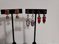 Three pairs of vintage non-pierced earrings displayed on black earring stands against plain background. Left pair amber-colored square clip-ons, center pair purple art deco screw-back, right pair red clip-on earrings with floral details.