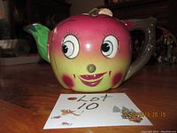 Front view showing apple-shaped teapot with smiling face and green spout, brown handle.