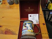 Photo of ceramic Christmas Carol tankard from 1975 inside original box with red lining, showing part of the embossed and painted design and 'LOT 2' sign.