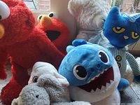 Photo showing various plush toys clustered together including two red Elmo characters, a blue Baby Shark plush, Pete the Cat plush, white teddy bear and other neutral color plush animals.