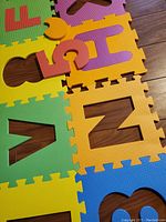 Close-up of several colorful foam mats with removable letter and number pieces, showing letters F, U, N, H, and 5 in purple, red, yellow, and orange colors arranged on a wooden floor.