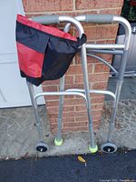 Front view of aluminum folding walker showing 2-button release handles, wheels on front legs, tennis ball covers on back legs, and attached red/black pouch.