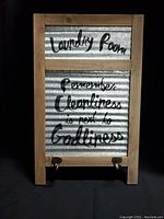 Front view of wooden washboard with corrugated metal panel and printed message 'Laundry Room Remember Cleanliness is next to Godliness'. Two metal hooks attached at bottom.