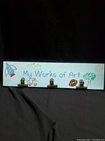 Full view of the wooden wall hanger painted light blue, showing four metal clips and the phrase 'My Works of Art' with child-themed illustrations.