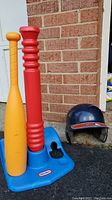 Full view of the Little Tikes T-ball set showing the blue base with two plastic bats - one red and one yellow - inserted into the base, and the navy-blue helmet placed beside the set.