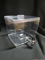 Clear plastic square beverage dispenser with attached spigot and removable lid, photographed against black background