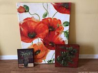Three pieces of floral wall art shown together against a wall: a large canvas painting featuring red poppies, a small framed print of a potted plant, and a red relief plaque with floral design.