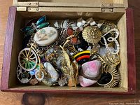 Photo of a wooden box filled with assorted brooches and pins showing various colors, shapes, and materials.