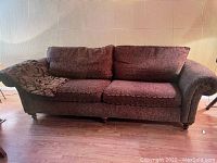 Brown fabric sofa couch with rolled arms, two seat cushions and two back cushions, wooden legs, and a dark patterned throw over the left armrest shown against a light wall and wood floor.